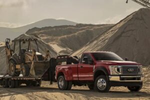 Red Ford Super Duty truck towing a flatbed trailer loaded with a backhoe on a construction site — representing AR (Air Brake Restricted) License training for medium-duty vehicles over 4,500 kg and under 11,000 kg — NEXTGEN Driver Training & Compliance.