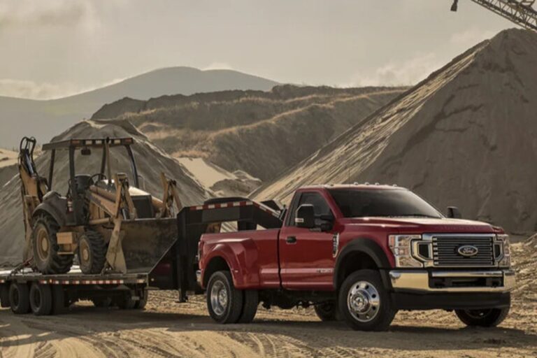 Red Ford Super Duty truck towing a flatbed trailer loaded with a backhoe on a construction site — representing AR (Air Brake Restricted) License training for medium-duty vehicles over 4,500 kg and under 11,000 kg — NEXTGEN Driver Training & Compliance.