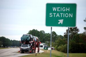 A car carrier truck loaded with multiple vehicles enters a highway weigh station, passing a green roadside sign marked “WEIGH STATION” with an arrow pointing toward the inspection area.