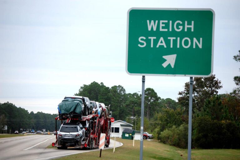A car carrier truck loaded with multiple vehicles enters a highway weigh station, passing a green roadside sign marked “WEIGH STATION” with an arrow pointing toward the inspection area.