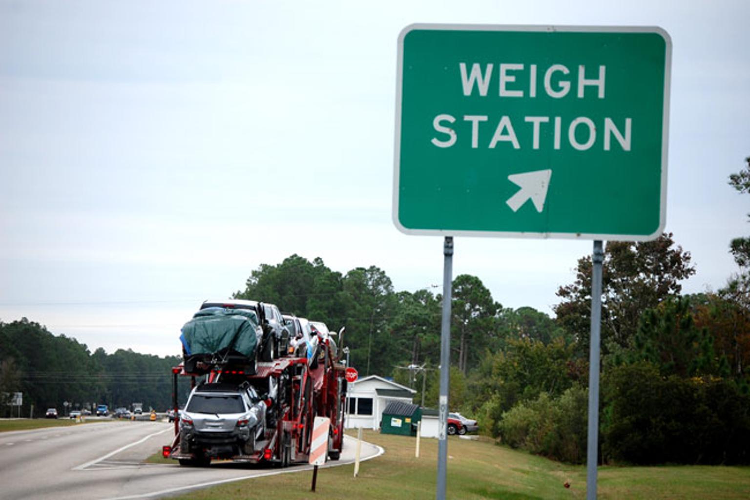 A car carrier truck loaded with multiple vehicles enters a highway weigh station, passing a green roadside sign marked “WEIGH STATION” with an arrow pointing toward the inspection area.