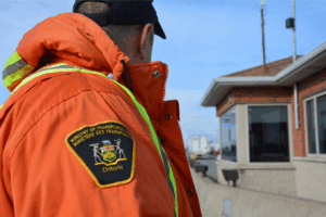 Ontario Ministry of Transportation (MTO) enforcement officer wearing a high-visibility orange jacket with reflective stripes stands near a roadside inspection station on a clear day.